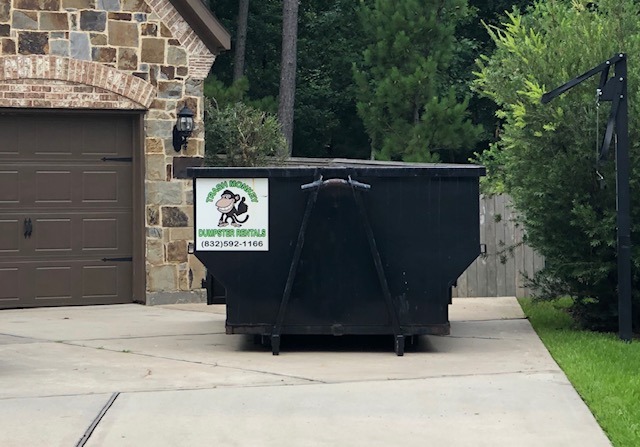 Black dumpster located in a driveway, featuring a logo and contact information for a dumpster rental service. Lush trees and a stone garage visible in the background.