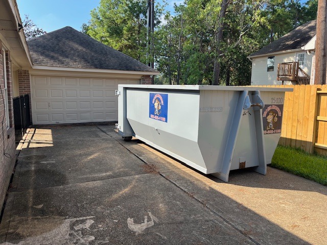 A large gray dumpster is positioned in a driveway beside a garage, surrounded by trees and residential buildings, indicating ongoing home renovations or clean-up.