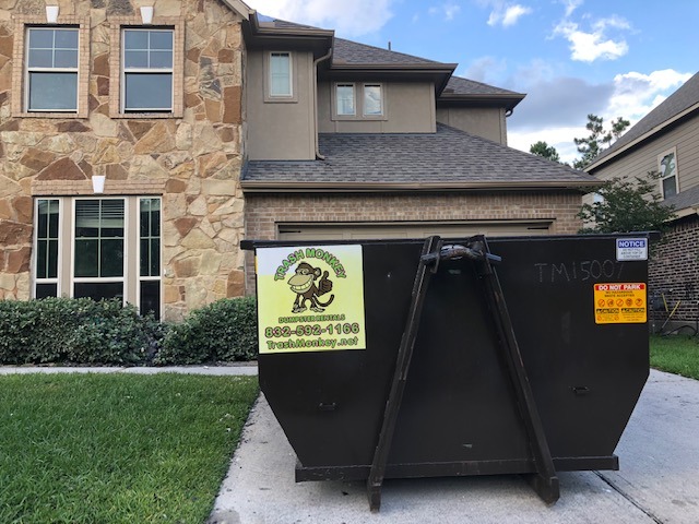 A large black dumpster with a colorful "Trash Monkey" logo sits in front of a modern home, indicating ongoing cleanup or renovation activities.