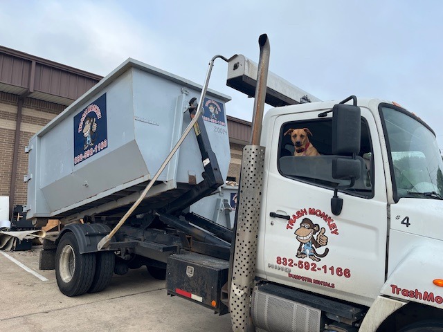 A trash truck with a light blue dumpster is parked. A friendly dog sits in the driver's seat, showcasing the company "Trash Monkey" branding.