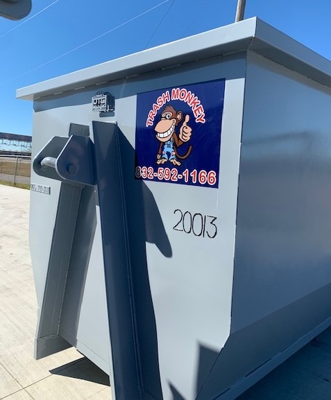Gray dumpster with a "Trash Monkey" logo featuring a cartoon monkey and contact number, set against a clear blue sky.