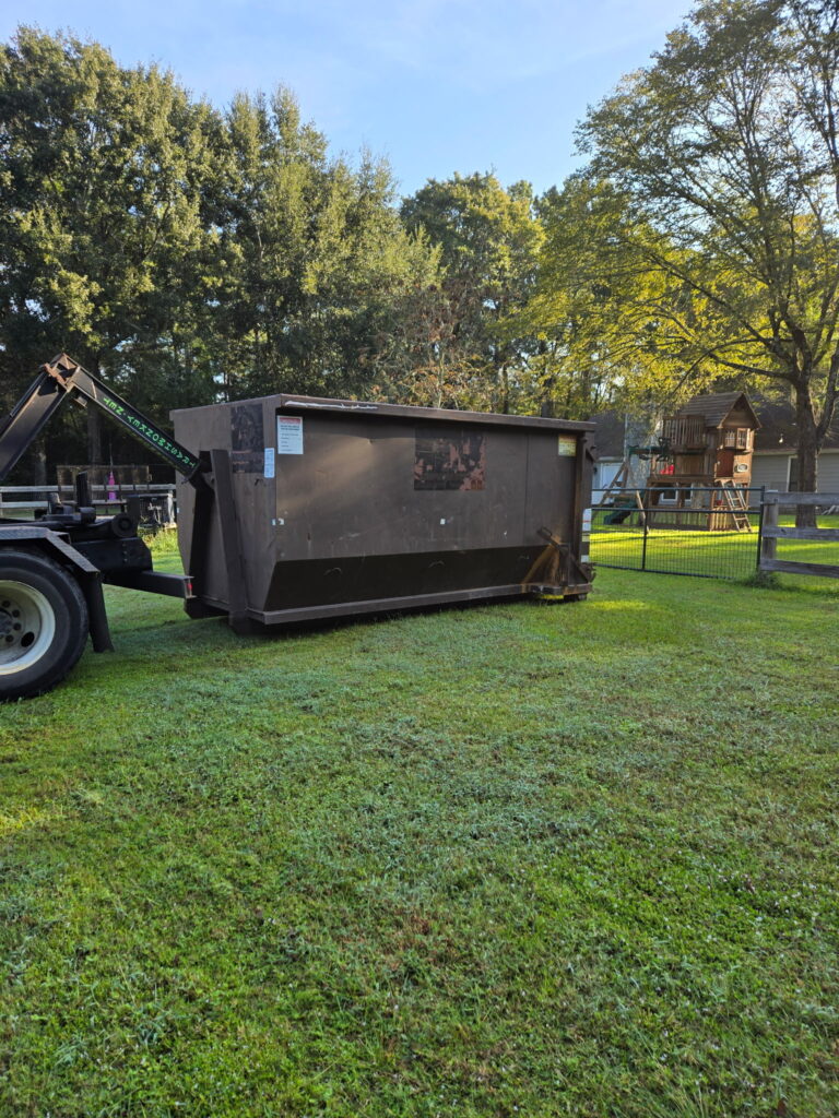 A large, brown dumpster sits on grass in a yard, with trees and a playset visible in the background, indicating a cleanup or renovation effort.