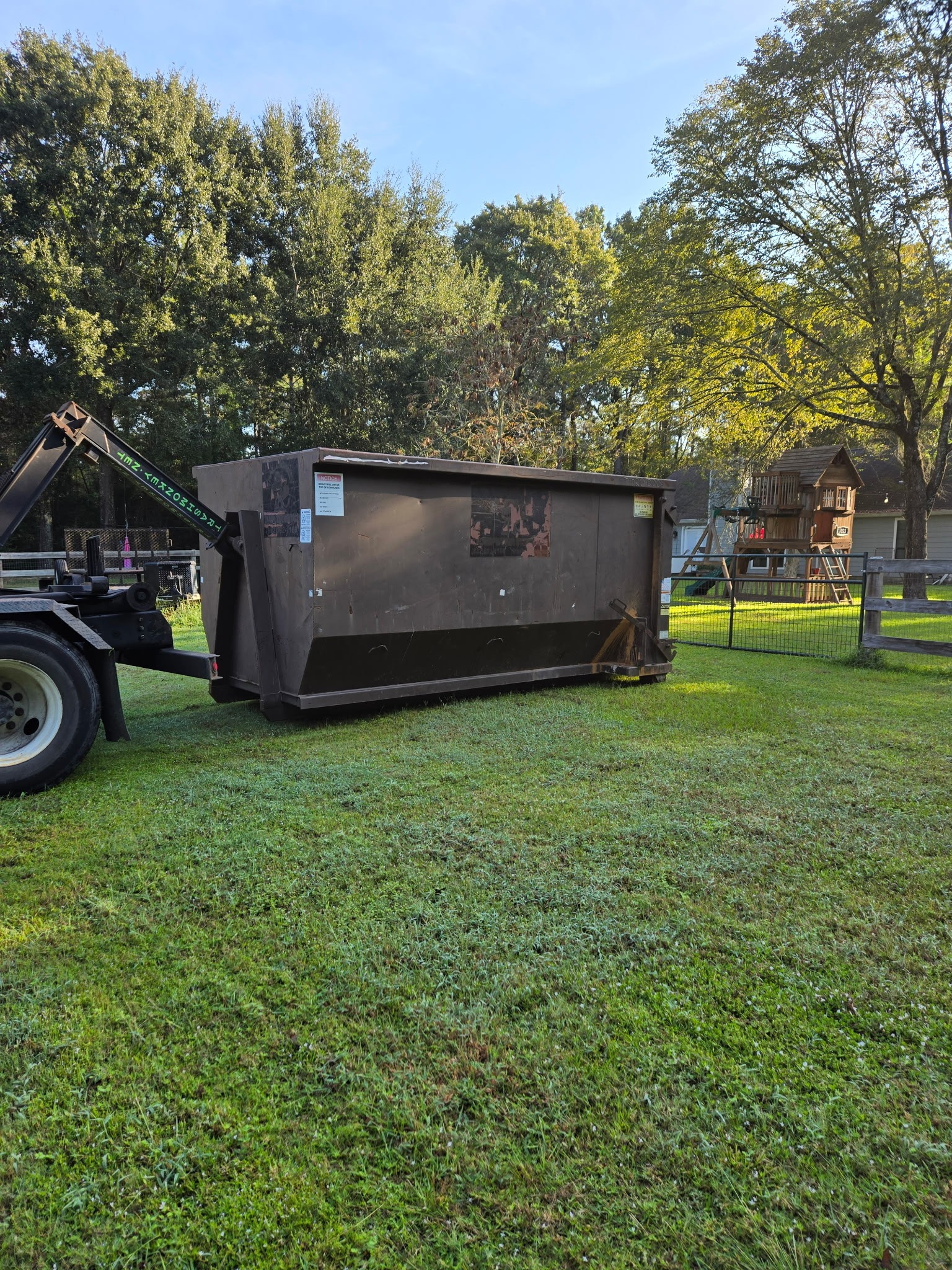 A large, brown dumpster sits on grass in a yard, with trees and a playset visible in the background, indicating a cleanup or renovation effort.