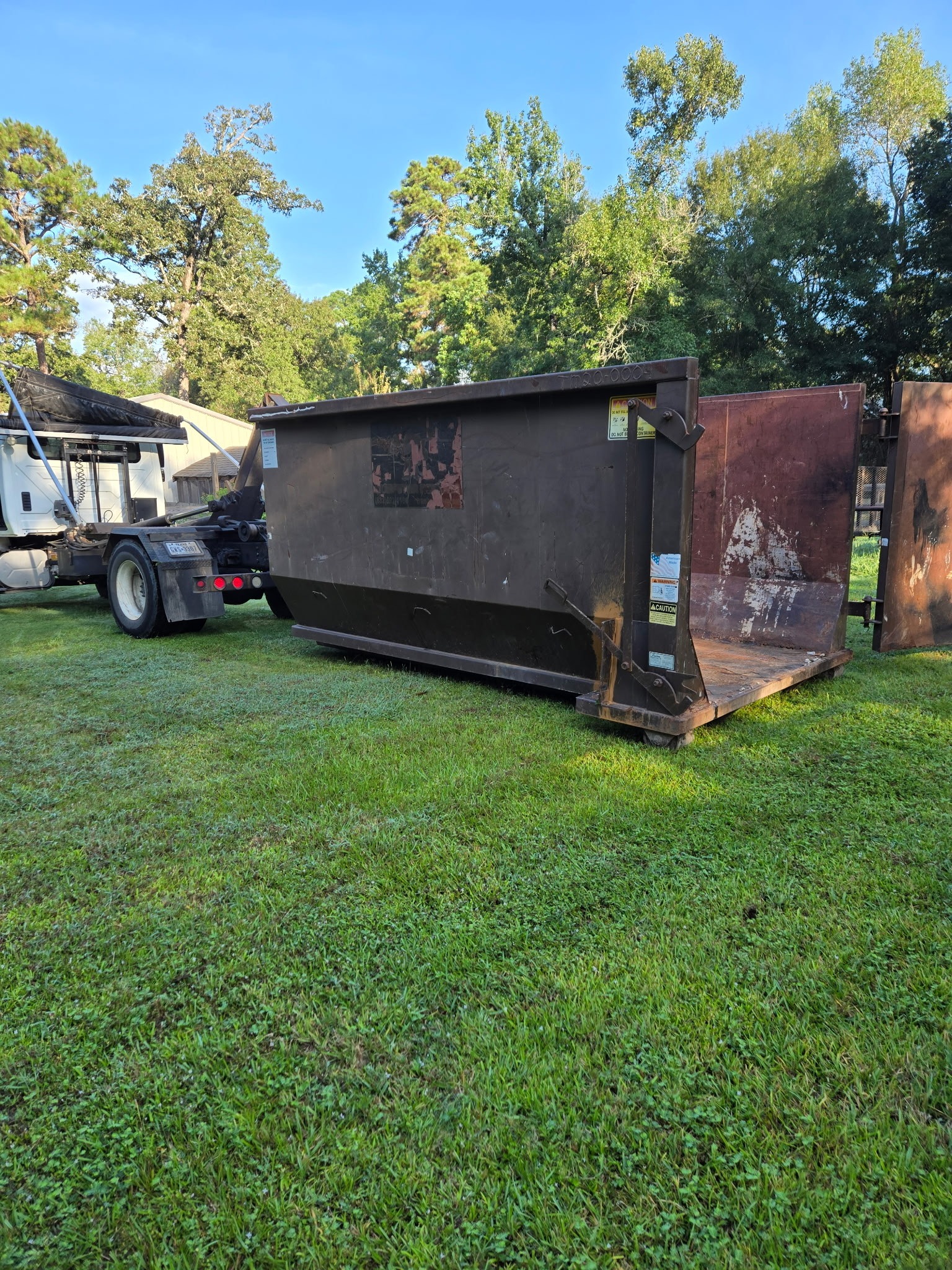 A large, dark brown dumpster sits on green grass, with a truck nearby for waste removal. The scene depicts a residential cleanup setup.